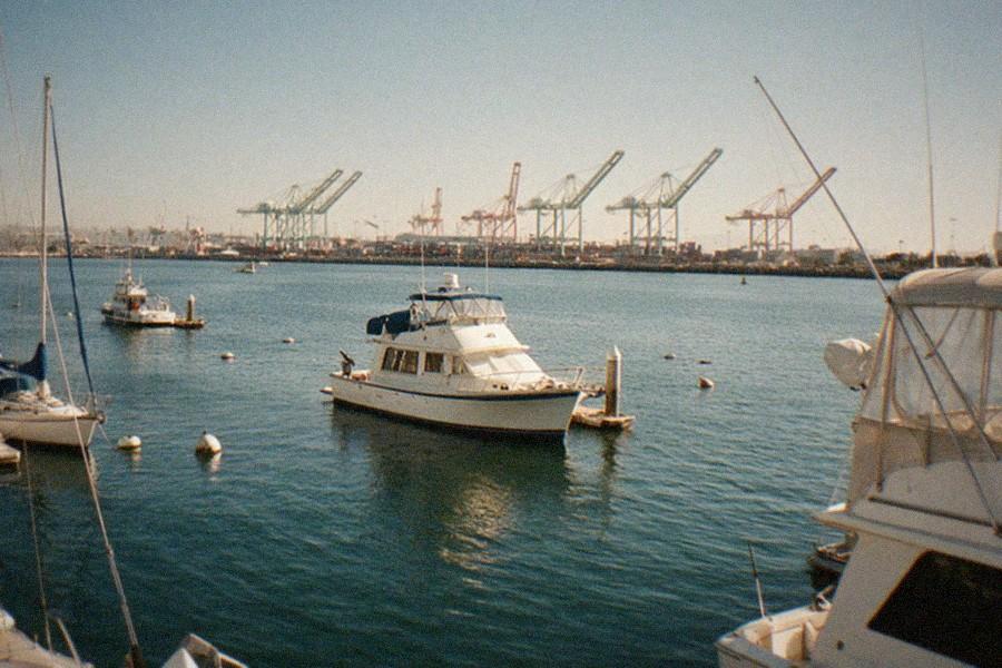 A view of a harbor channel with a gray battleship moored along the dock and cranes on the far shore